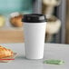 A white Choice paper hot cup with a black lid next to a croissant on a table.