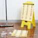 A yellow caution sign on the floor with a Rubbermaid Over-The-Spill medium absorbent pad tablet and mop.