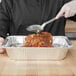 A person using a Vollrath stainless steel basting spoon to pour sauce on a piece of meat in a tin pan.