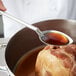 A person holding a Vollrath stainless steel basting spoon over a pot of meat with brown liquid.