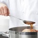 A hand using a Vollrath stainless steel spoon to pour brown sauce into a pan.