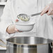 A person in a white chef's coat using a Vollrath slotted basting spoon to stir a silver pot.