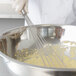A person in a white chef's uniform using a Vollrath stainless steel whisk to mix dough in a bowl.