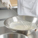 A person in a white coat using a Vollrath stainless steel whisk to mix white liquid in a bowl.