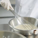 A person in white gloves using a Vollrath stainless steel French whisk to stir a bowl of liquid.