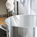 A person in a white coat using a Vollrath stainless steel whisk to mix food in a large pot.