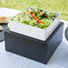 A salad on a table in a square container on a white Cal-Mil Midnight Cube Riser.