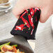 A hand holding a black fabric pot handle cover with chili peppers on it over a pan of food.