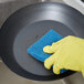 A person wearing a yellow glove using a 3M blue non-stick cookware cleaning pad to clean a pan.