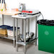 An Advance Tabco stainless steel filler table with a backsplash and undershelf in a kitchen with a green garbage can and a table.