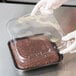 A person in gloves holding a Solut clear plastic lid over a brown cake in a plastic container.