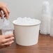 A hand pouring ice from a Lavex white paper ice bucket into a plastic cup.