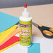 A Creativity Street Chenille Kraft White Liquid Tacky Glue bottle next to scissors and colored paper.
