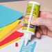A hand holding a white bottle of Creativity Street Chenille Kraft Tacky Glue over a colorful paper.