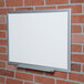 A Universal deluxe white melamine dry erase board with aluminum frame on a brick wall.