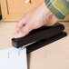 A person's hand using a black Universal full strip stapler to staple a piece of paper on a black surface.
