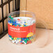 A jar of Universal plastic push pins in assorted rainbow colors on a table.