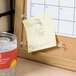 A close up of a container of Universal clear plastic push pins with a cork board in the background with a sticky note pinned to it.