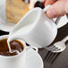 A person pouring cream from a white Tuxton creamer into a cup of coffee.