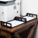 A black Universal plastic desk tray holding a black and white paper on a wooden table with other trays of papers next to a printer.