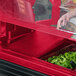 A hand places food in a red Cambro Versa Well cover on a salad bar counter.