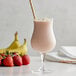 A glass of pink Monin Strawberry Banana smoothie with a yellow and white striped straw on a table with a strawberry and a banana.