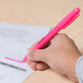 A hand holding a Universal Fluorescent Pink Chisel Tip Highlighter over a piece of paper.