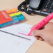 A hand using a Universal fluorescent pink highlighter to mark on a paper with a group of colorful markers.