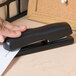 A person's hand using a black Bostitch ergonomic desktop stapler to staple a piece of paper.