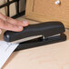 A person's hand using a Bostitch black ergonomic desktop stapler on a table.