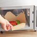 A hand reaching out to a Kraft microwavable folded paper take-out box filled with a burger, broccoli, and an apple.