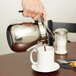 A hand holding a Thunder Group stainless steel coffee decanter with a black handle pouring coffee into a white mug.