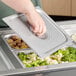 A hand holding a Vigor stainless steel steam table pan cover over a tray of food.
