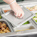 A hand in a plastic glove holding a Vigor stainless steel steam table pan cover over a tray of food.