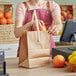 A woman in a pink apron using a brown paper bag with a handle to put oranges on a table.
