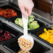 A hand holding a white plastic spoon over a container of food on a salad bar.