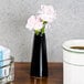 An American Metalcraft black tower vase with pink flowers on a wooden surface next to a cup of coffee.