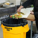 A person in a black chef's uniform putting food into a yellow Rubbermaid BRUTE trash can.