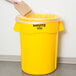 A person putting a cardboard box in a yellow Rubbermaid trash can.