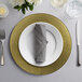 A place setting with a 10 Strawberry Street gold rim glass charger plate, silverware, and a napkin on a table.