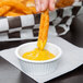A person dipping a french fry into a bowl of yellow sauce using a white fluted plastic ramekin.