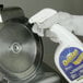 A person wearing gloves uses a spray bottle to clean a circular meat slicer blade.