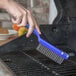 A hand with a blue plastic handle using a Carlisle Sparta Wire Grill Brush to clean a grill.