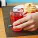 A hand holding a Cambro clear plastic tumbler filled with red juice on a table with a stack of pancakes.
