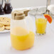 A Tablecraft 48 oz. dispenser of orange juice on a table next to a glass of orange juice.