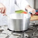 A person in a white coat stirring food in a Vollrath aluminum sauce pan.