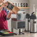 A person pouring coffee beans from a brown bag into a Bunn double hopper coffee grinder on a counter in a coffee shop.