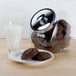 A glass jar of chocolate cookies on a plate and a glass of milk on a table.