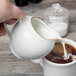 A hand holding a 10 Strawberry Street Gold Line porcelain creamer pouring milk into a white cup of coffee.