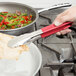 A hand using Vollrath stainless steel tongs with a red Kool Touch handle to serve food from a pan on a stove.